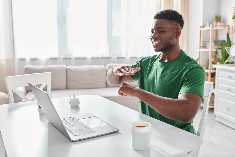 Man in a green shirt smiling and gesturing with his hands while sitting at a table with a laptop, coffee mug, and notepad in a bright room.